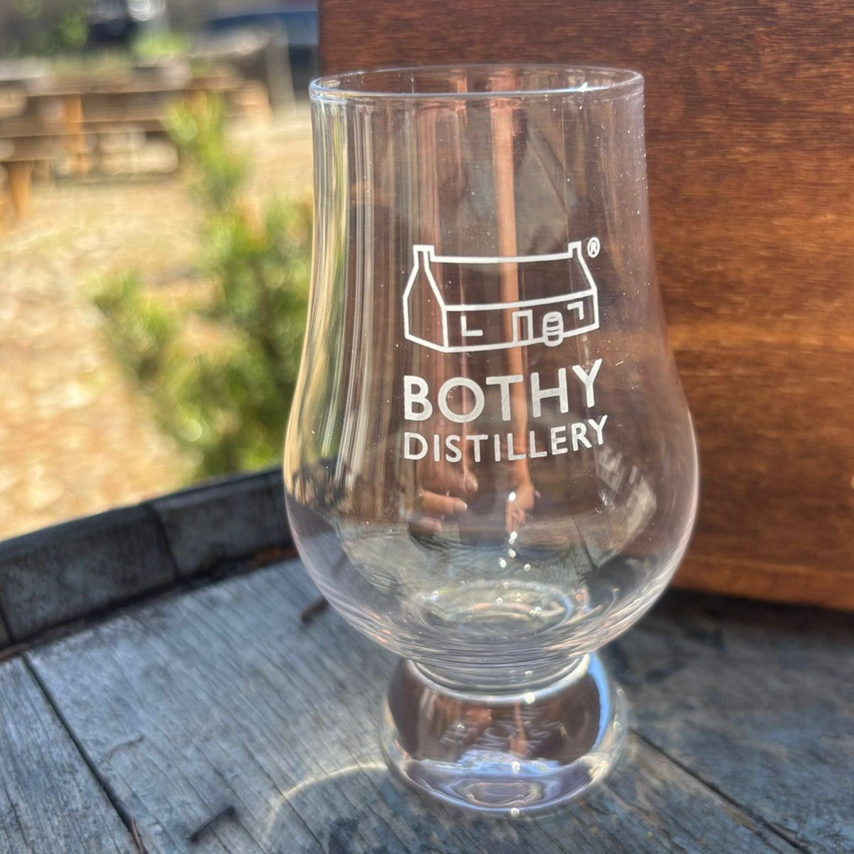 A clear Glencairn glass with 'Bothy Distillery' printed on it, featuring a narrow neck and flared bowl, placed on a wooden surface.
