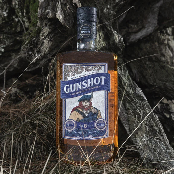 A bottle of Gunshot Blended Scotch Whisky placed against a backdrop of rocks and grass, with a label featuring an illustration of a man's face.