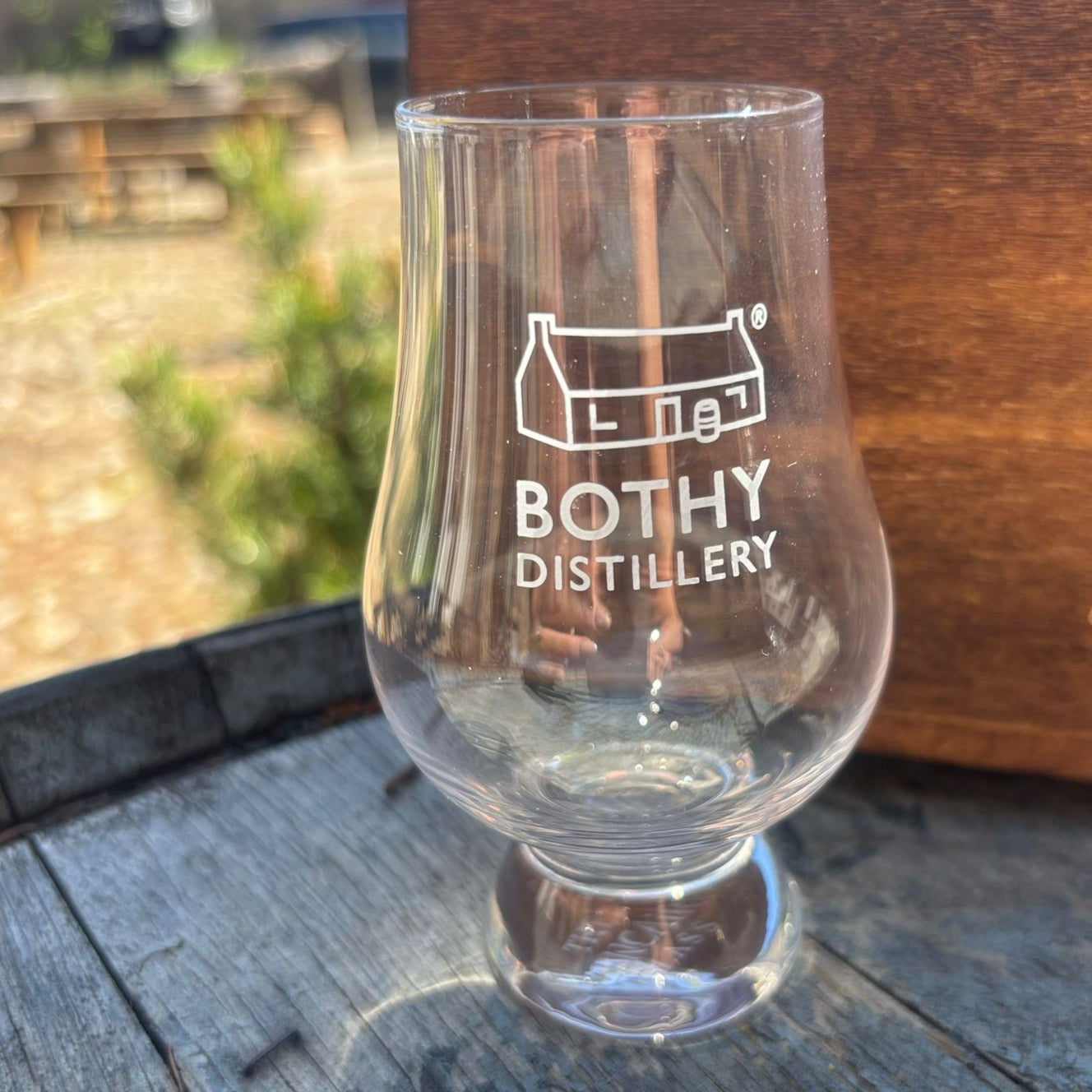 A clear Glencairn glass with 'Bothy Distillery' printed on it, featuring a narrow neck and flared bowl, placed on a wooden surface.