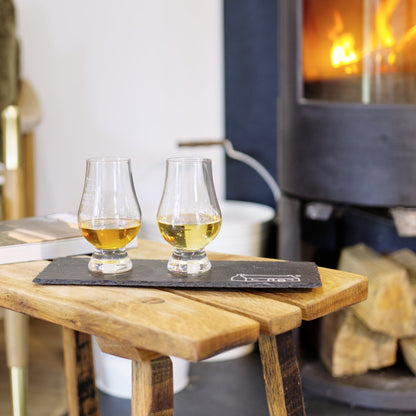 Two glasses of whiskey on a wooden table in front of a burning fireplace.