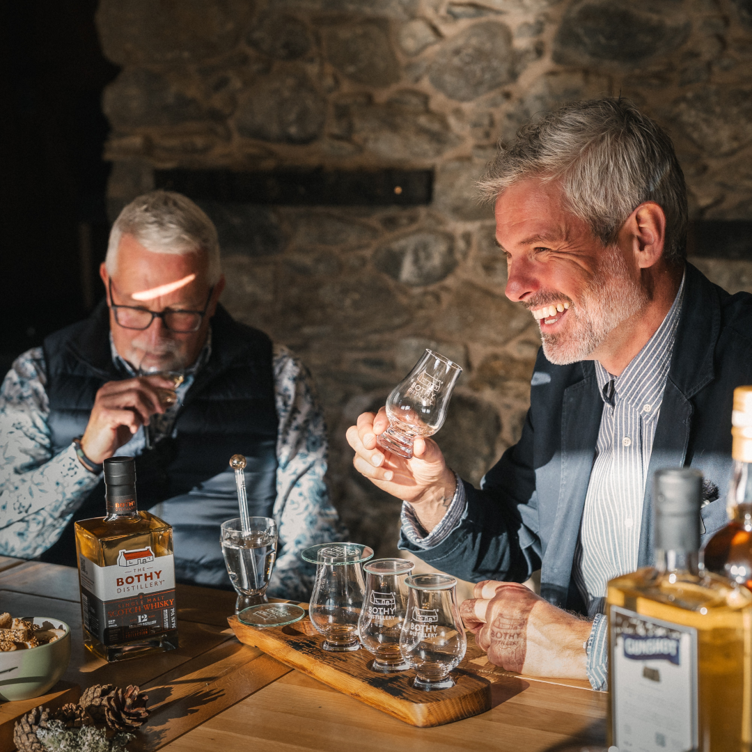 Two men enjoying a bottle of Bothy whisky in a cozy setting with stone walls.