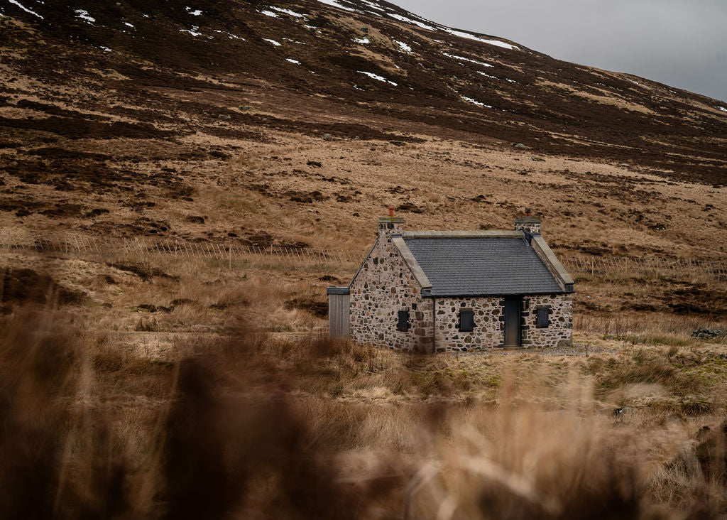 Stone cottage in a mountainous landscape with dry grass and a cloudy sky.