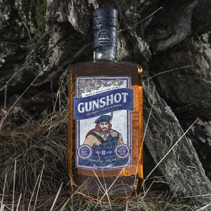 A bottle of Gunshot Blended Scotch Whisky placed against a backdrop of rocks and grass, with a label featuring an illustration of a man's face.