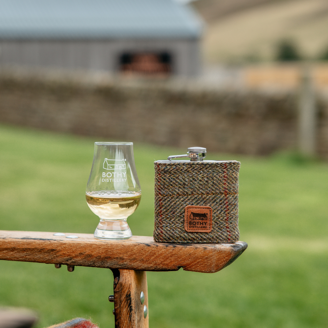 Harris Tweed hip flask and glass with 'Bottle & Board' branding on a wooden surface outdoors.