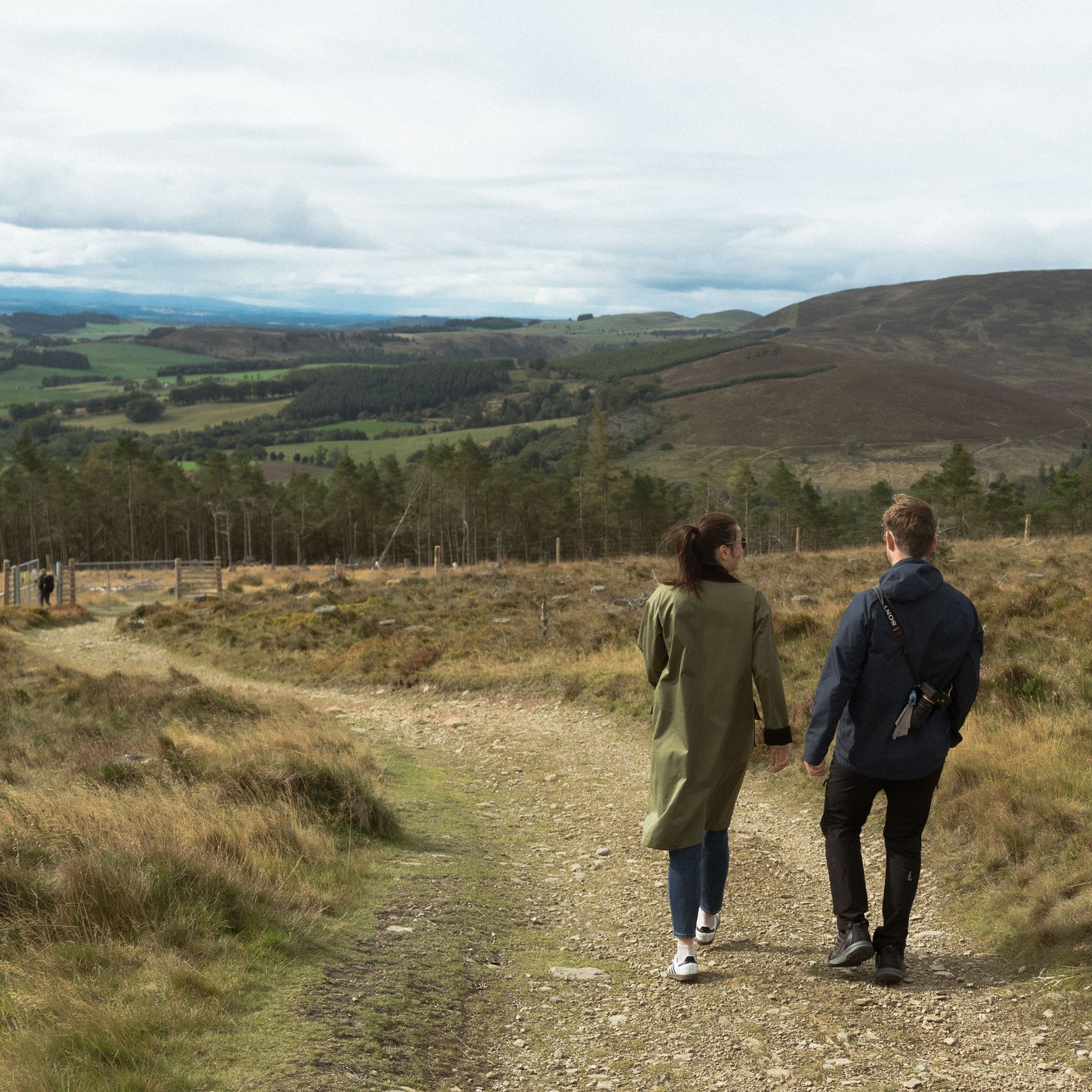 Two people walking along a dirt path in a scenic landscape with hills and trees.