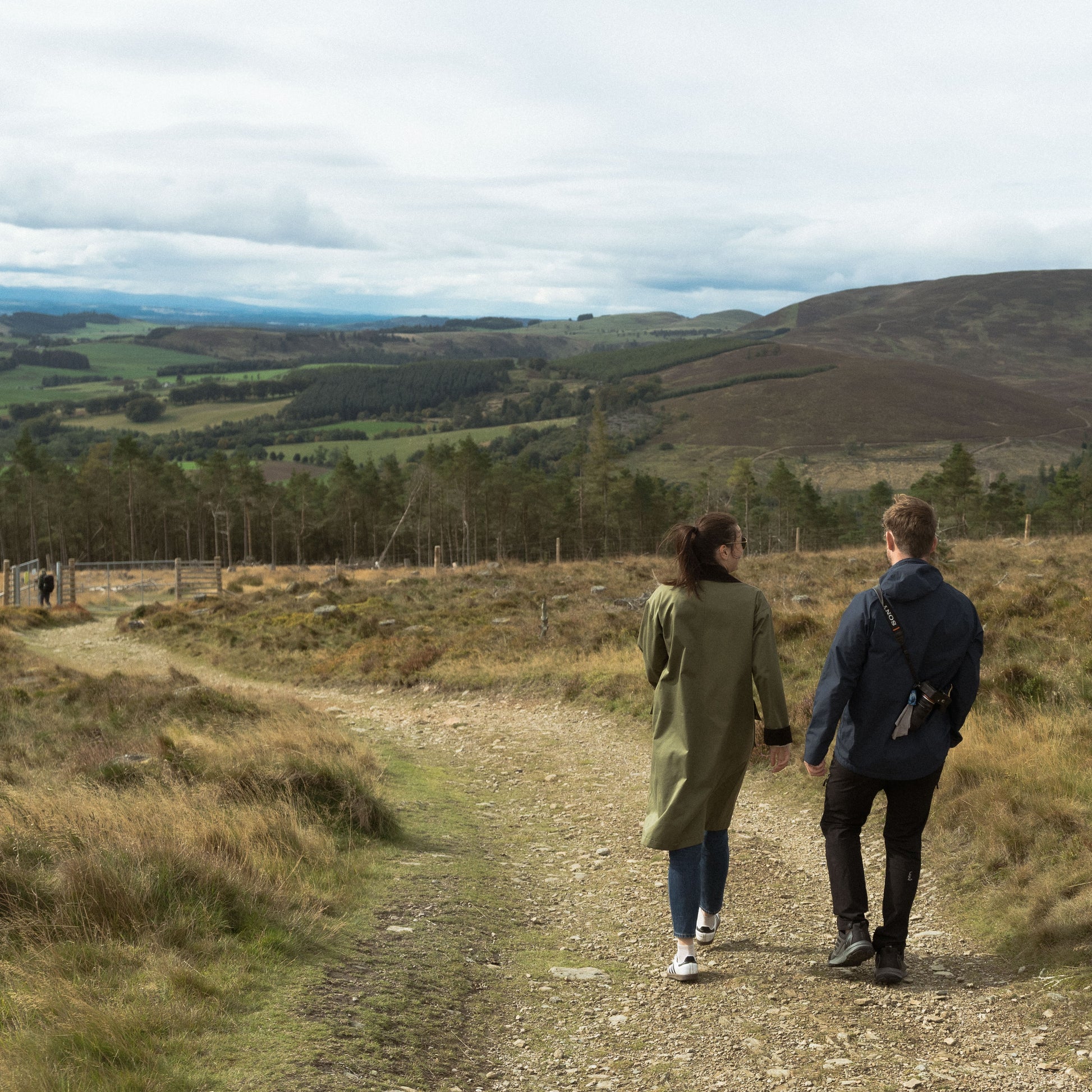 Two people walking along a dirt path in a scenic landscape with hills and trees.