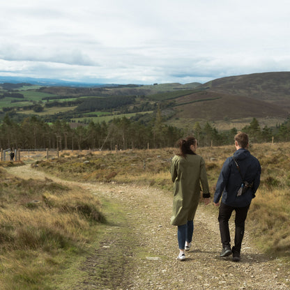 Two people walking along a dirt path in a scenic landscape with hills and trees.